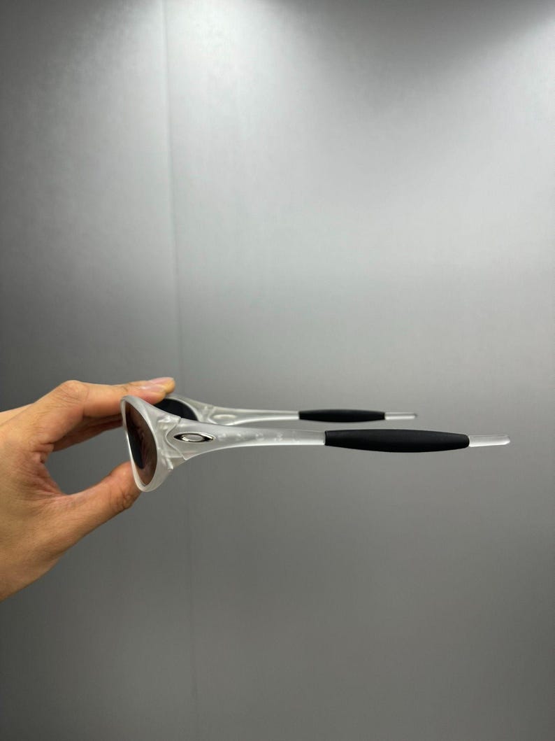 May include: Clear-framed sunglasses with dark lenses and black temples. The sunglasses are held up, showcasing the design and shape. The frame has a sleek, modern design, suitable for outdoor activities.
