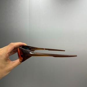 Puede incluir: Gafas de sol marrones y naranjas con un dise&ntilde;o elegante y moderno. Las lentes son de un naranja vibrante, que contrasta con las monturas y las patillas marrones. Las gafas de sol se sostienen, mostrando su forma y estilo. Un peque&ntilde;o logotipo es visible en la montura.