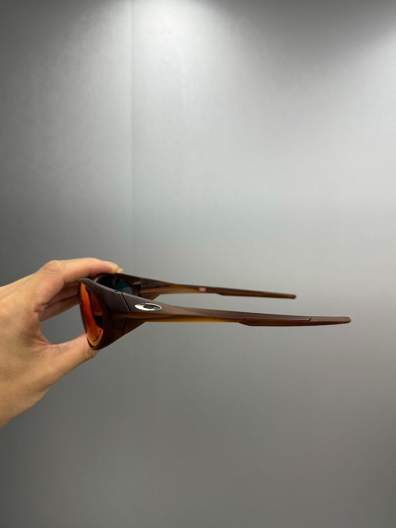 Puede incluir: Gafas de sol marrones y naranjas con un dise&ntilde;o elegante y moderno. Las lentes son de un naranja vibrante, que contrasta con las monturas y las patillas marrones. Las gafas de sol se sostienen, mostrando su forma y estilo. Un peque&ntilde;o logotipo es visible en la montura.
