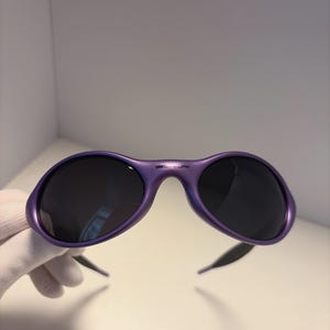 K&ouml;nnte beinhalten: Violette Sonnenbrille mit dunklen Gl&auml;sern. Die Sonnenbrille hat ein geschwungenes, umlaufendes Design und eine gl&auml;nzende Oberfl&auml;che. Der Markenname ist auf der Oberseite des Rahmens aufgedruckt. Die B&uuml;gel sind schwarz.