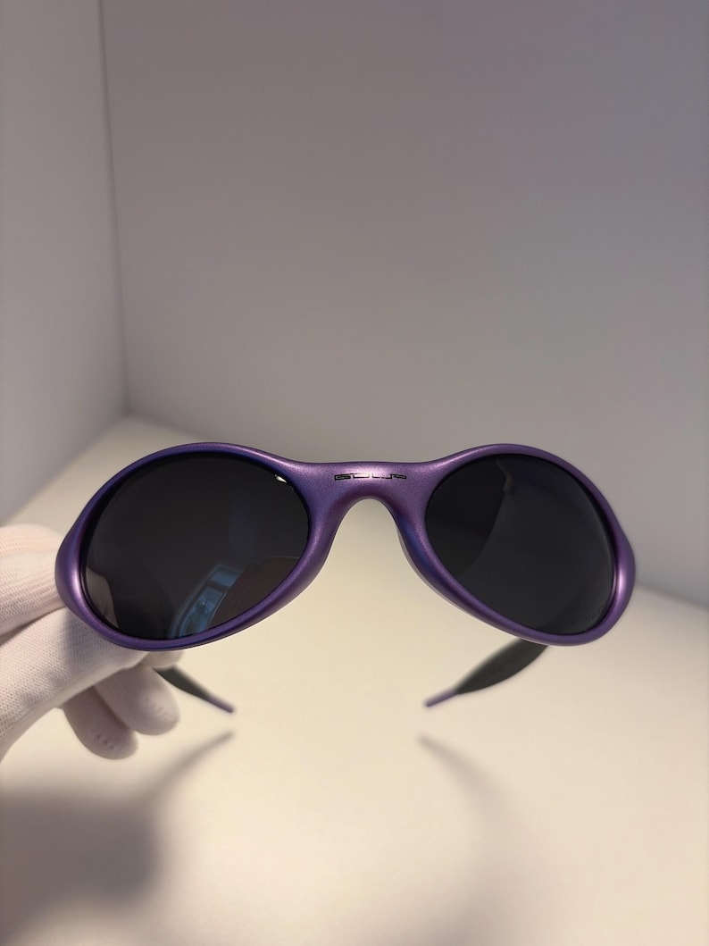 K&ouml;nnte beinhalten: Violette Sonnenbrille mit dunklen Gl&auml;sern. Die Sonnenbrille hat ein geschwungenes, umlaufendes Design und eine gl&auml;nzende Oberfl&auml;che. Der Markenname ist auf der Oberseite des Rahmens aufgedruckt. Die B&uuml;gel sind schwarz.