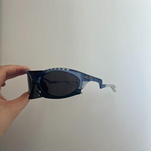May include: Blue and black sunglasses with a distinctive, angular design. The sunglasses have dark lenses and a light blue frame with a unique shape. The arms are a light grey colour. The Oakley logo is visible on the frame.