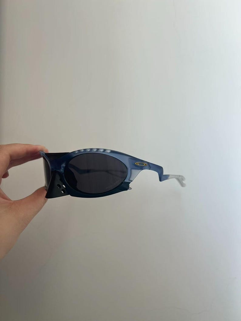 May include: Blue and black sunglasses with a distinctive, angular design. The sunglasses have dark lenses and a light blue frame with a unique shape. The arms are a light grey colour. The Oakley logo is visible on the frame.