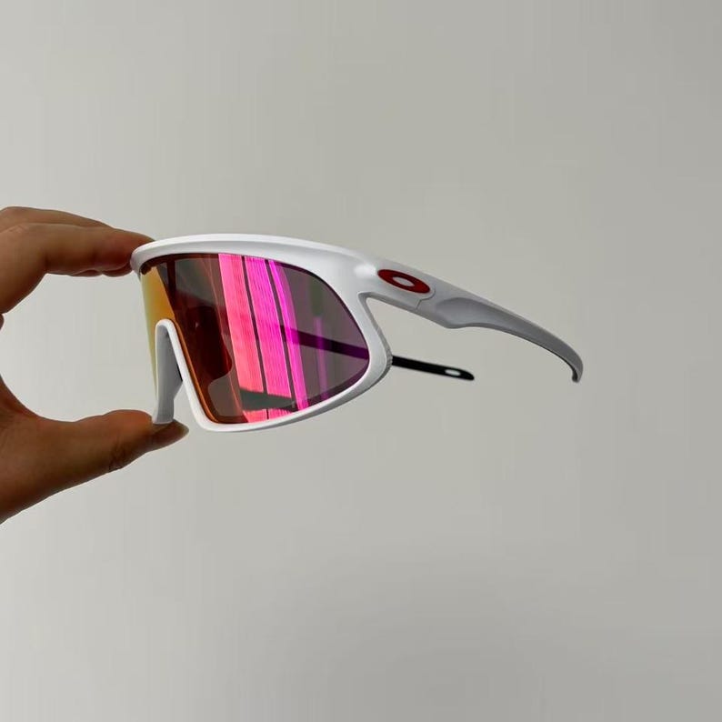 May include: White sports sunglasses with a red logo on the temple. The lenses have a gradient of colors, including pink and orange. The sunglasses are held by a hand.