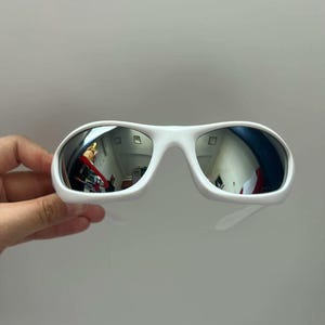 K&ouml;nnte beinhalten: Wei&szlig;e Sonnenbrille mit verspiegelten Gl&auml;sern. Die Sonnenbrille hat einen dicken, abgerundeten Rahmen und B&uuml;gel. Die Gl&auml;ser spiegeln die Umgebung wider. Die Sonnenbrille wird von einer Hand vor einem schlichten Hintergrund gehalten.