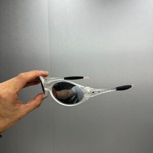 May include: Clear-framed sunglasses with mirrored lenses. The sunglasses have black accents on the temples and nose bridge. The lenses reflect the surrounding environment. The frame is held by a hand against a gray background.