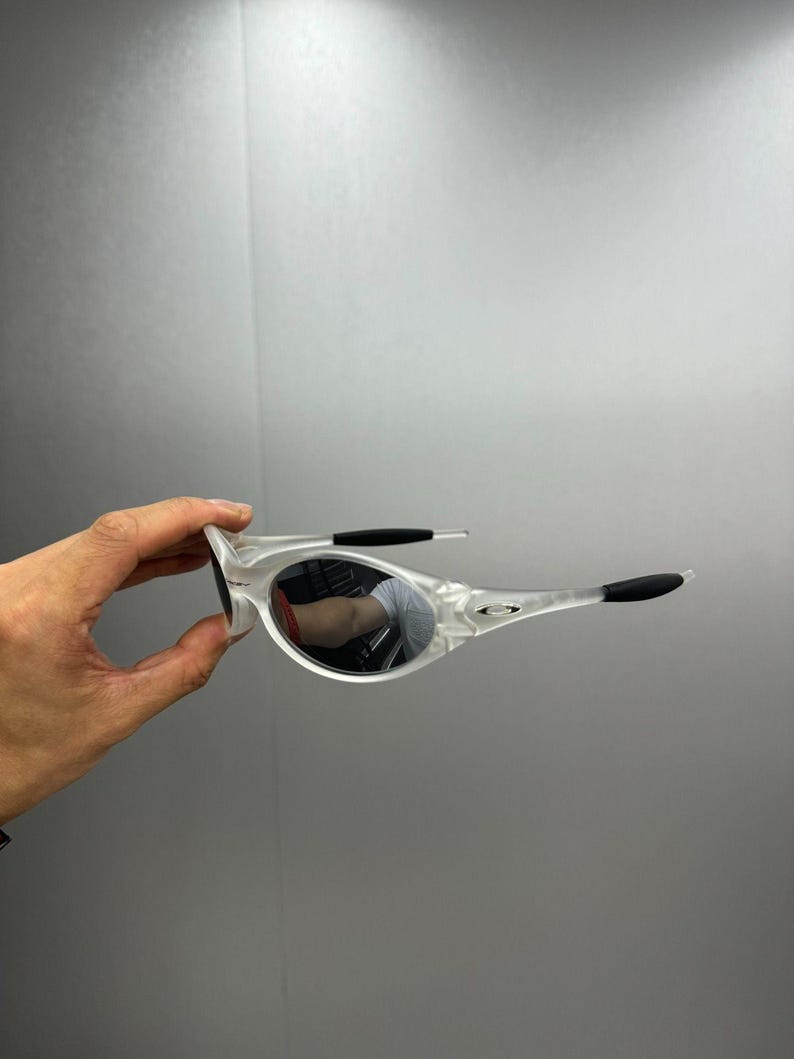 May include: Clear-framed sunglasses with mirrored lenses. The sunglasses have black accents on the temples and nose bridge. The lenses reflect the surrounding environment. The frame is held by a hand against a gray background.