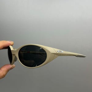 May include: Tan-colored sunglasses with oval lenses and a wraparound design. The sunglasses have a sleek, modern aesthetic, with the brand name visible on the arm. The lenses are dark, providing contrast to the frame.