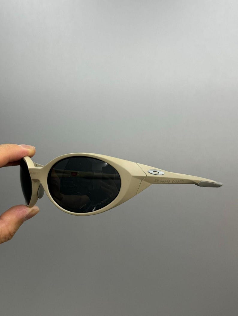 May include: Tan-colored sunglasses with oval lenses and a wraparound design. The sunglasses have a sleek, modern aesthetic, with the brand name visible on the arm. The lenses are dark, providing contrast to the frame.