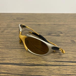 May include: Gold and silver-toned sunglasses with brown lenses. The sunglasses have a curved design and a black rubber piece on the temple. The frame is a combination of gold and silver colors.