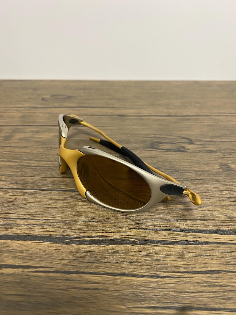 May include: Gold and silver-toned sunglasses with brown lenses. The sunglasses have a curved design and a black rubber piece on the temple. The frame is a combination of gold and silver colors.