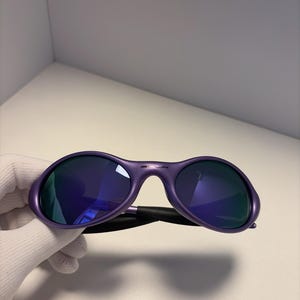 May include: Purple sunglasses with dark blue lenses. The sunglasses have a rounded frame and a black arm. The lenses reflect the surrounding environment. The sunglasses are held by a gloved hand.