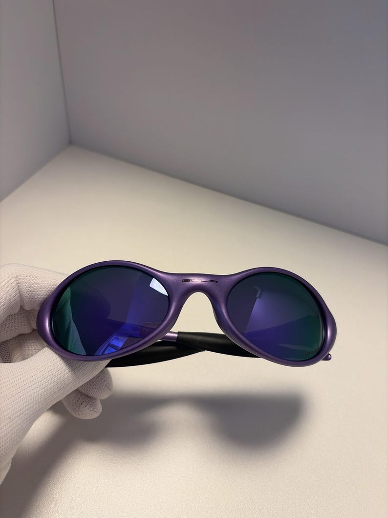 May include: Purple sunglasses with dark blue lenses. The sunglasses have a rounded frame and a black arm. The lenses reflect the surrounding environment. The sunglasses are held by a gloved hand.