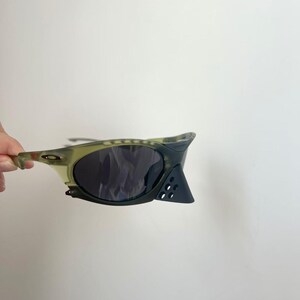May include: A pair of olive green and black sunglasses with dark lenses. The sunglasses have a unique design with a curved frame and a triangular accent on the side. The lenses are dark, and the frame has a camouflage pattern.