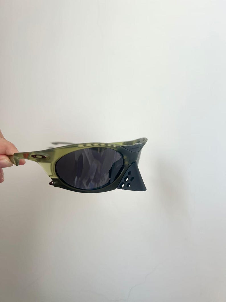 May include: A pair of olive green and black sunglasses with dark lenses. The sunglasses have a unique design with a curved frame and a triangular accent on the side. The lenses are dark, and the frame has a camouflage pattern.