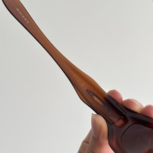May include: Close-up of brown sunglasses with a curved frame. The sunglasses have a dark brown tint and the arms are a lighter shade of brown. The text "05-013 60 20" is printed on the arm.