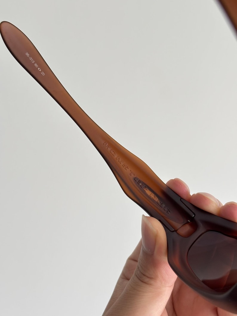 May include: Close-up of brown sunglasses with a curved frame. The sunglasses have a dark brown tint and the arms are a lighter shade of brown. The text "05-013 60 20" is printed on the arm.