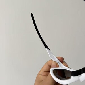 May include: White sunglasses with black accents. The sunglasses have a sleek, modern design with a white frame and dark lenses. The arms are white with black tips. The sunglasses are held by a hand.