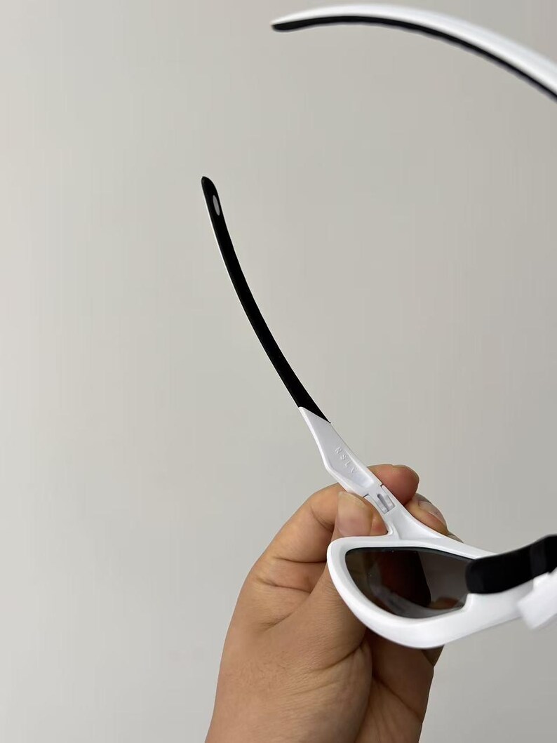 May include: White sunglasses with black accents. The sunglasses have a sleek, modern design with a white frame and dark lenses. The arms are white with black tips. The sunglasses are held by a hand.