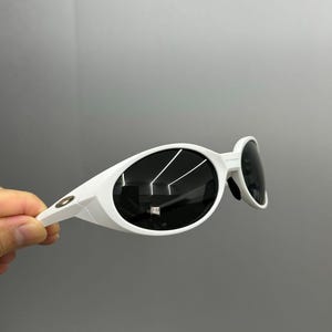 K&ouml;nnte beinhalten: Wei&szlig;e, ovale Sonnenbrille mit dunklen Gl&auml;sern. Die Sonnenbrille hat ein modernes Design mit einem dicken, wei&szlig;en Rahmen. Die Gl&auml;ser reflektieren das Licht, und die B&uuml;gel sind ebenfalls wei&szlig;.