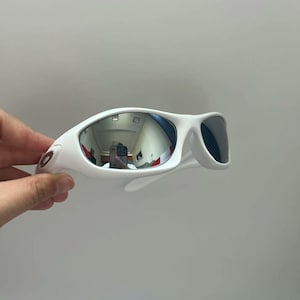 K&ouml;nnte beinhalten: Wei&szlig;e Sonnenbrille mit sportlichem Design. Die verspiegelten Gl&auml;ser reflektieren die Umgebung. Der Rahmen ist einfarbig wei&szlig;, und die B&uuml;gel sind f&uuml;r einen sicheren Sitz gebogen. Die Sonnenbrille hat eine moderne, sportliche &Auml;sthetik.