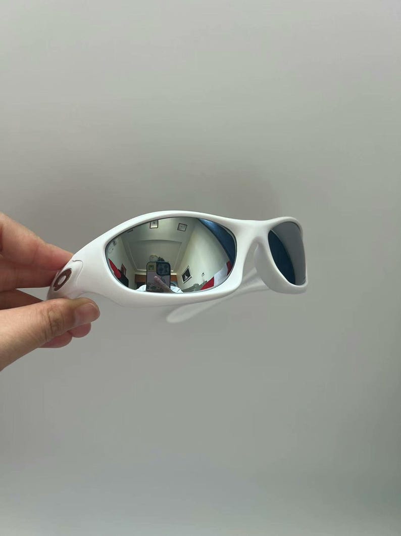 K&ouml;nnte beinhalten: Wei&szlig;e Sonnenbrille mit sportlichem Design. Die verspiegelten Gl&auml;ser reflektieren die Umgebung. Der Rahmen ist einfarbig wei&szlig;, und die B&uuml;gel sind f&uuml;r einen sicheren Sitz gebogen. Die Sonnenbrille hat eine moderne, sportliche &Auml;sthetik.