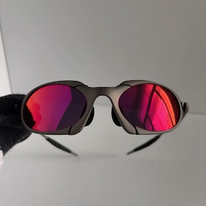 Op de afbeelding: Een zonnebril met een donkergrijs montuur en felrode glazen. De glazen reflecteren licht, waardoor een heldere, kleurrijke uitstraling ontstaat. De zonnebril heeft een strak, modern design.