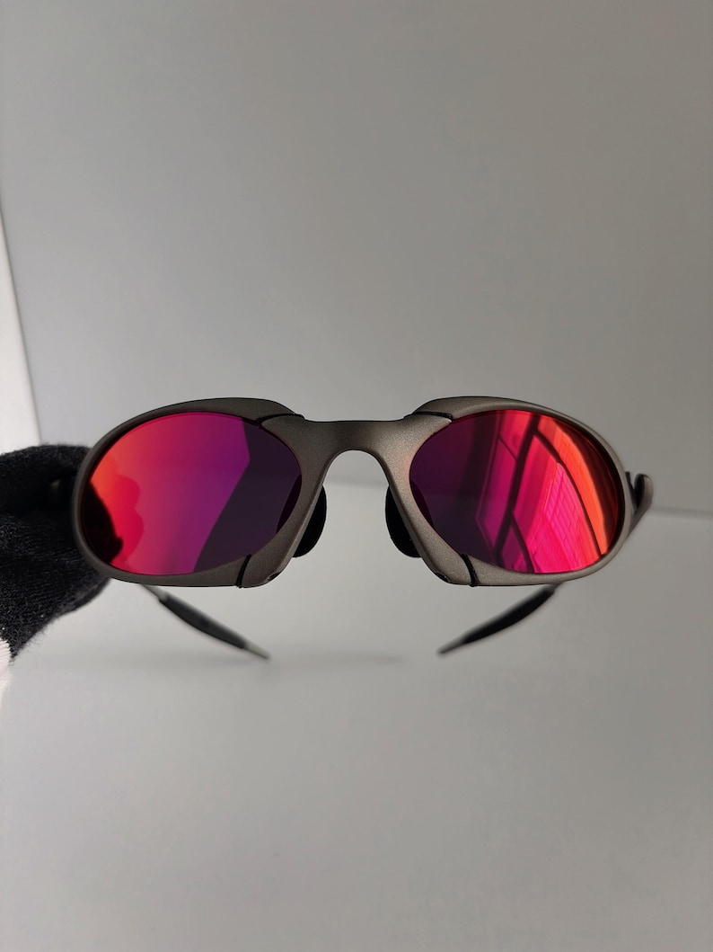 May include: A pair of sunglasses with a dark gray frame and vibrant red lenses. The lenses reflect light, creating a bright, colorful appearance. The sunglasses have a sleek, modern design.