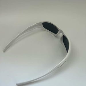 K&ouml;nnte beinhalten: Wei&szlig;e Sonnenbrille mit schwarzen Gl&auml;sern. Die Sonnenbrille hat ein sportliches Design mit gebogenem Rahmen und B&uuml;geln. Die Gl&auml;ser sind rechteckig und bieten vollen Schutz. Geeignet f&uuml;r Outdoor-Aktivit&auml;ten.