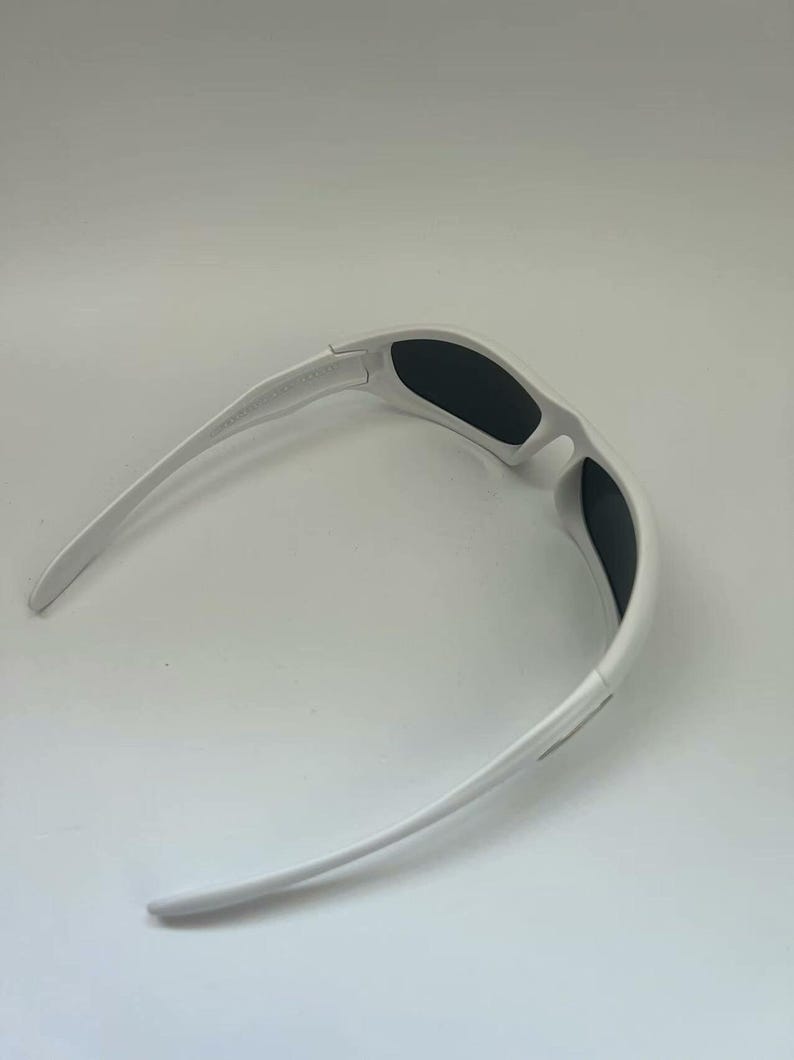 K&ouml;nnte beinhalten: Wei&szlig;e Sonnenbrille mit schwarzen Gl&auml;sern. Die Sonnenbrille hat ein sportliches Design mit gebogenem Rahmen und B&uuml;geln. Die Gl&auml;ser sind rechteckig und bieten vollen Schutz. Geeignet f&uuml;r Outdoor-Aktivit&auml;ten.