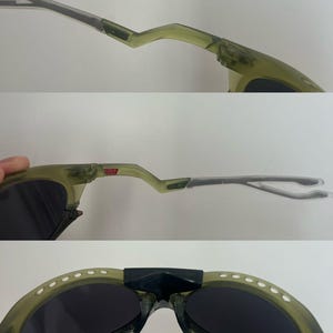 May include: Olive green sunglasses with dark lenses. The frames have a unique, angular design with a bridge and temples. The sunglasses have a small "USA" label on the frame.