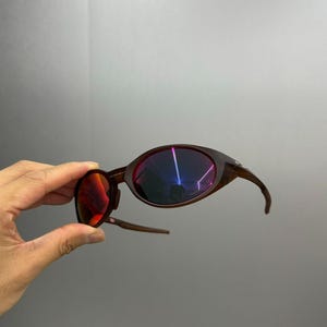 Puede incluir: Gafas de sol marrones con una forma ovalada inusual y patillas curvas. Las lentes tienen un revestimiento reflectante multicolor. Las gafas de sol se sostienen sobre un fondo gris.