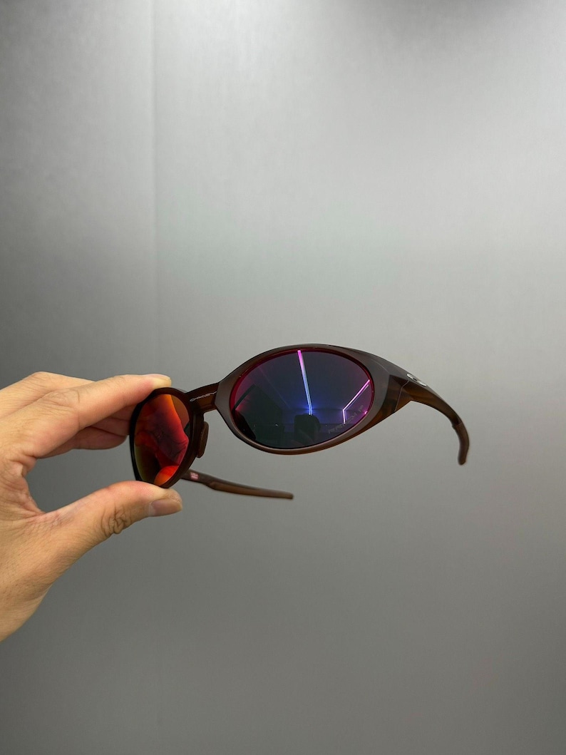 Puede incluir: Gafas de sol marrones con una forma ovalada inusual y patillas curvas. Las lentes tienen un revestimiento reflectante multicolor. Las gafas de sol se sostienen sobre un fondo gris.