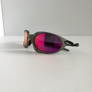 May include: A pair of gray sunglasses with vibrant red and purple lenses. The sunglasses have a sleek, modern design with a curved frame and a small, orange-tinted lens on the left side. The lenses reflect light, creating a gradient effect.