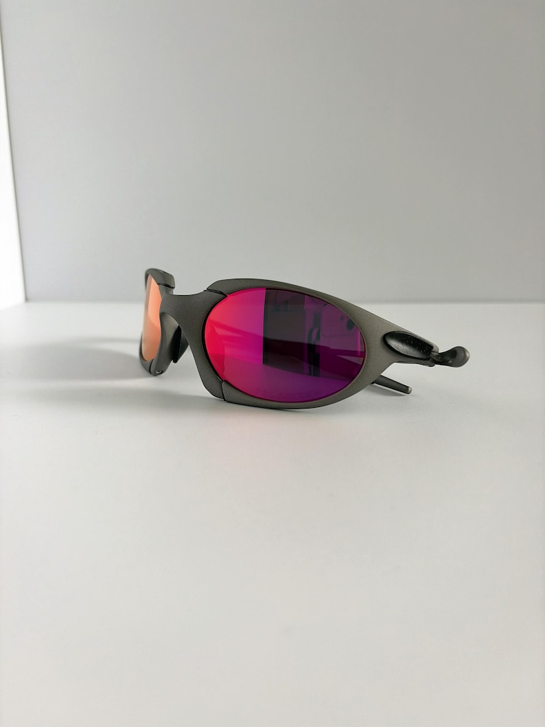 May include: A pair of gray sunglasses with vibrant red and purple lenses. The sunglasses have a sleek, modern design with a curved frame and a small, orange-tinted lens on the left side. The lenses reflect light, creating a gradient effect.