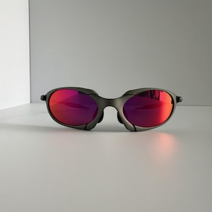 May include: A pair of gray sunglasses with vibrant red lenses. The sunglasses have a sleek, modern design with a curved frame and a bridge that sits low on the nose. The lenses reflect light, creating a bright, eye-catching appearance.