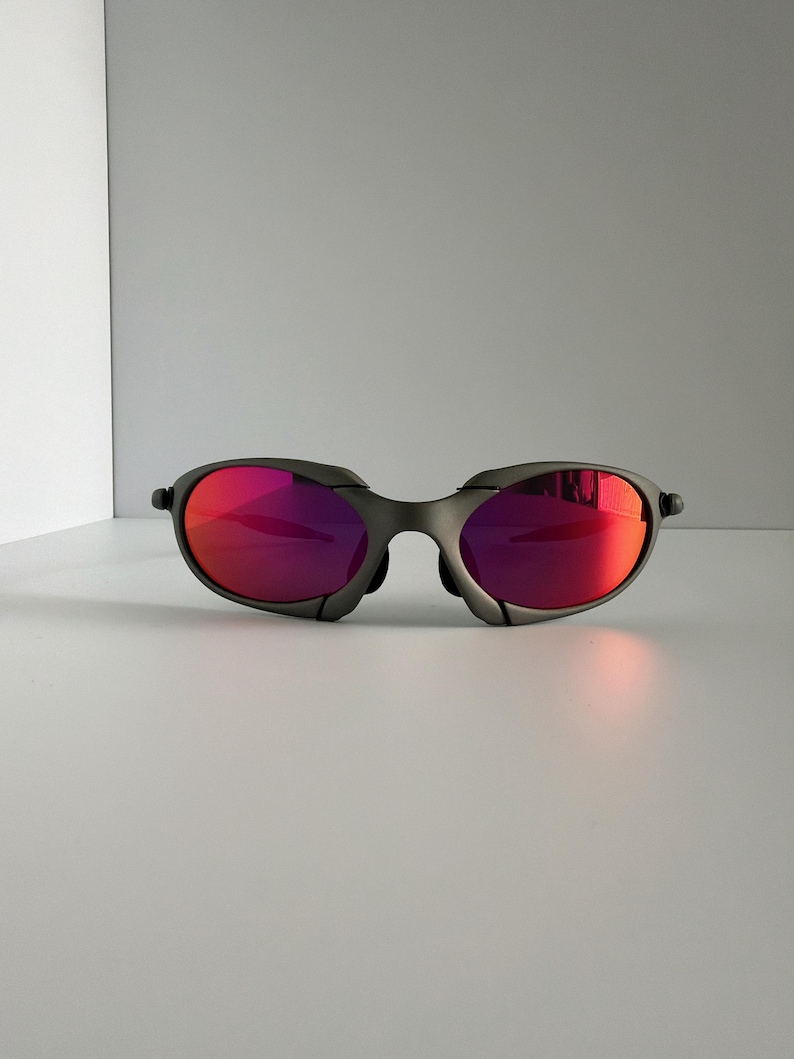 May include: A pair of gray sunglasses with vibrant red lenses. The sunglasses have a sleek, modern design with a curved frame and a bridge that sits low on the nose. The lenses reflect light, creating a bright, eye-catching appearance.