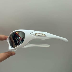 K&ouml;nnte beinhalten: Wei&szlig;e Sportsonnenbrille mit verspiegelten Gl&auml;sern. Die Sonnenbrille hat ein schlankes, modernes Design mit einem geschwungenen Rahmen und B&uuml;geln. Die Gl&auml;ser reflektieren die Umgebung. Das Oakley-Logo ist auf dem Rahmen sichtbar.