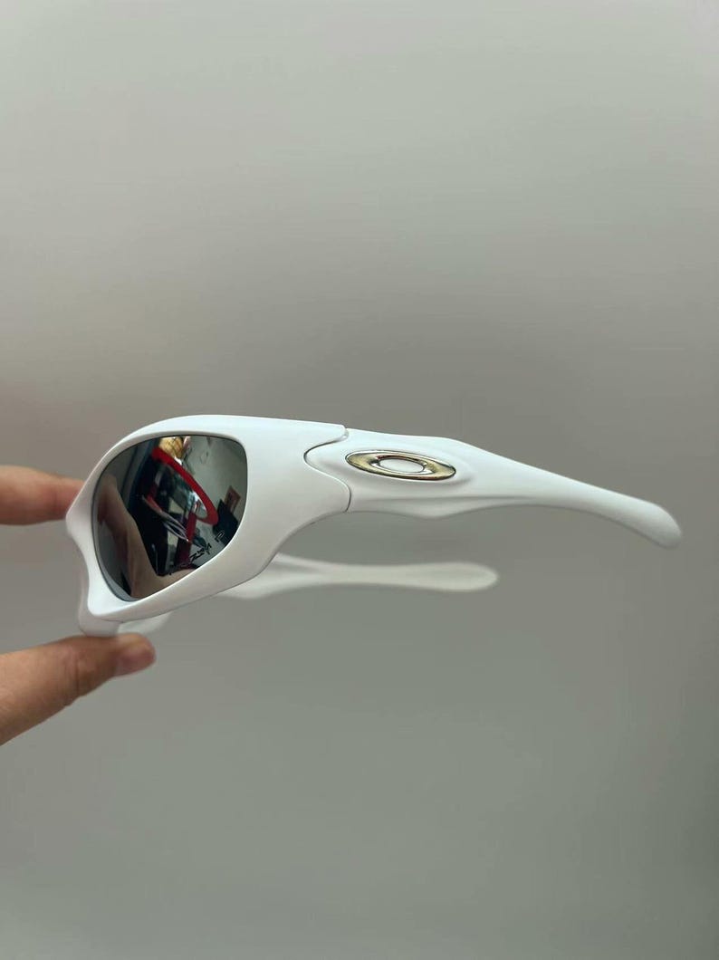 K&ouml;nnte beinhalten: Wei&szlig;e Sportsonnenbrille mit verspiegelten Gl&auml;sern. Die Sonnenbrille hat ein schlankes, modernes Design mit einem geschwungenen Rahmen und B&uuml;geln. Die Gl&auml;ser reflektieren die Umgebung. Das Oakley-Logo ist auf dem Rahmen sichtbar.