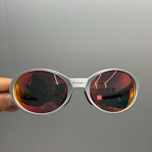 Puede incluir: Gafas de sol con montura plateada y lentes ovaladas con un tinte rojo-naranja. Las lentes reflejan el entorno. El nombre de la marca "OAKLEY" es visible en la montura. Las gafas se sostienen sobre un fondo gris.