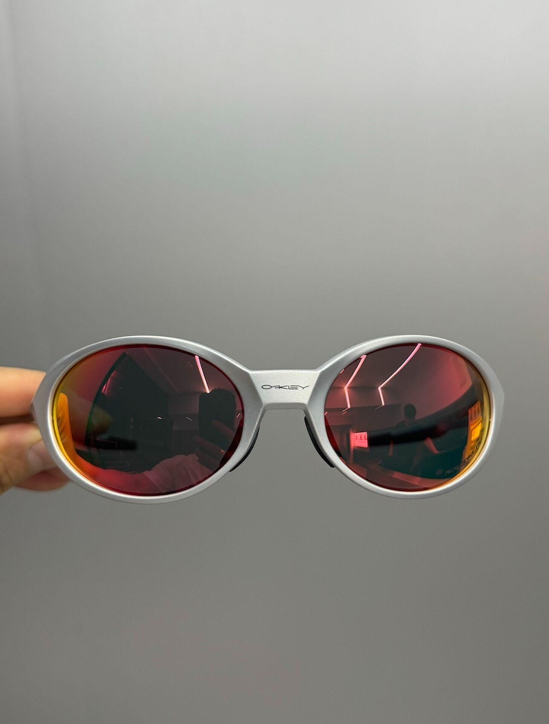 Puede incluir: Gafas de sol con montura plateada y lentes ovaladas con un tinte rojo-naranja. Las lentes reflejan el entorno. El nombre de la marca "OAKLEY" es visible en la montura. Las gafas se sostienen sobre un fondo gris.