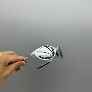 Pu&ograve; includere: Occhiali da sole bianchi con lenti a specchio. Gli occhiali hanno un design curvo e dettagli neri sulle aste. Le lenti riflettono l'ambiente circostante. Gli occhiali sono tenuti su uno sfondo grigio.