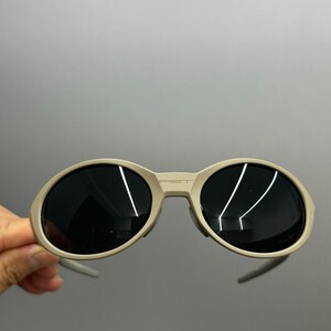 May include: Oval-shaped sunglasses with a light beige frame and dark lenses. The sunglasses have a bridge and arms. The lenses reflect light. The sunglasses are held against a gray background.
