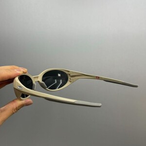 May include: Beige sunglasses with black lenses. The frames are a light tan color with gray arms. The sunglasses are held up against a gray background.