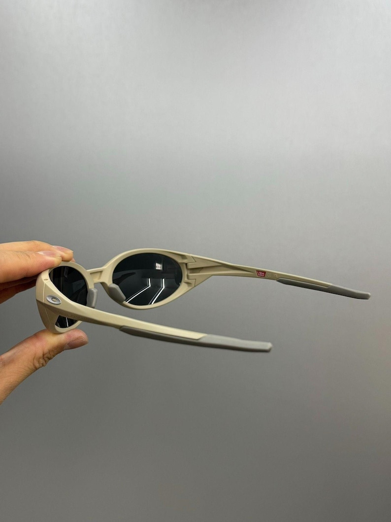May include: Beige sunglasses with black lenses. The frames are a light tan color with gray arms. The sunglasses are held up against a gray background.