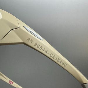 May include: Close-up of beige sunglasses with the text "RN 96548-CA35460" printed on the arm. The sunglasses have a modern design with a silver accent near the hinge and a red, white, and blue detail on the temple.