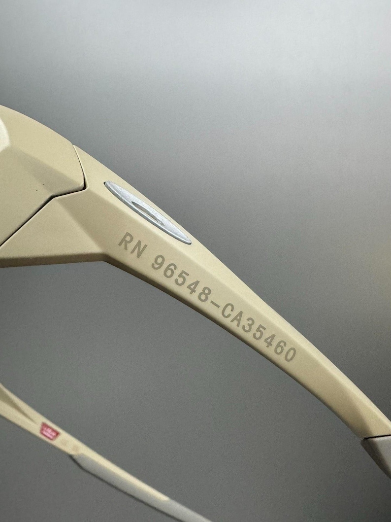 May include: Close-up of beige sunglasses with the text "RN 96548-CA35460" printed on the arm. The sunglasses have a modern design with a silver accent near the hinge and a red, white, and blue detail on the temple.