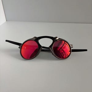 May include: A pair of sunglasses with a unique design. The lenses are a vibrant red, set in a dark metal frame with a black bridge and arms. The sunglasses have a distinctive, futuristic aesthetic.