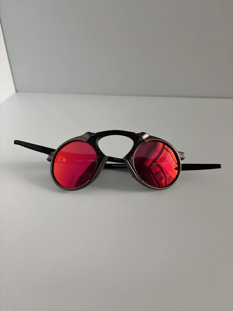 May include: A pair of sunglasses with a unique design. The lenses are a vibrant red, set in a dark metal frame with a black bridge and arms. The sunglasses have a distinctive, futuristic aesthetic.