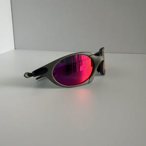 May include: A pair of gray sunglasses with vibrant pink lenses. The sunglasses have a sleek, modern design with a curved frame and a small, curved arm. The lenses reflect a bright pink hue, adding a pop of color to the overall look.